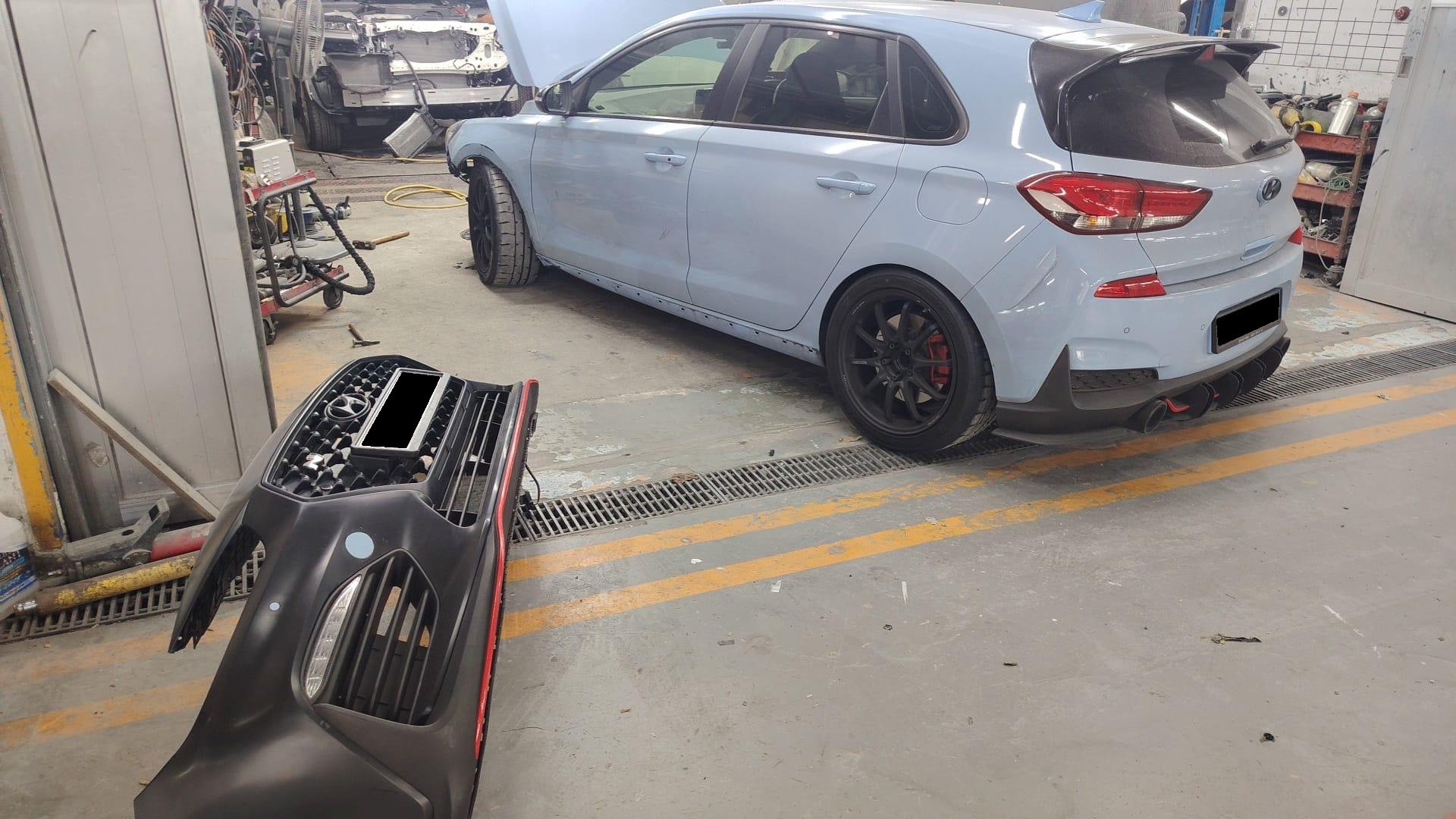Rear three-quarter in the body shop, front bumper lying in the foreground, plates obscured